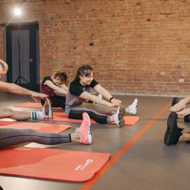 A group of diverse adults stretching on mats in a brick-walled fitness studio.